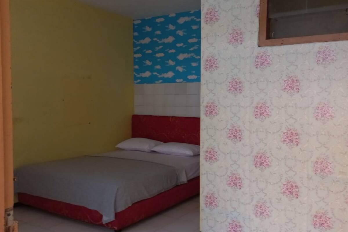 Un dormitori amb un panell de capçal vermell, un llit amb roba de llit gris, una paret groga, una paret blava amb dibuixos de núvols i una paret amb motius florals.