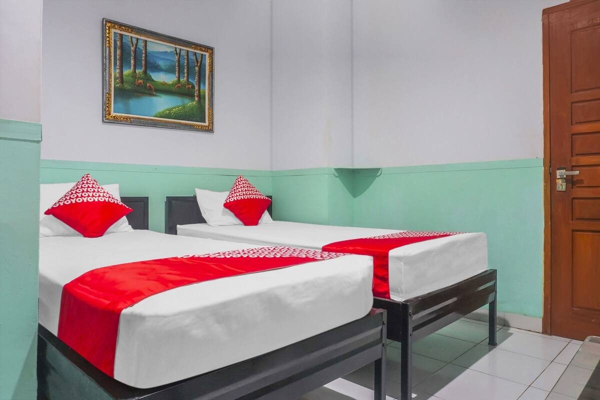 Une chambre avec deux lits, chacun agrémenté de draps blancs et d’oreillers rouges, un tableau encadré au mur et une porte en bois.