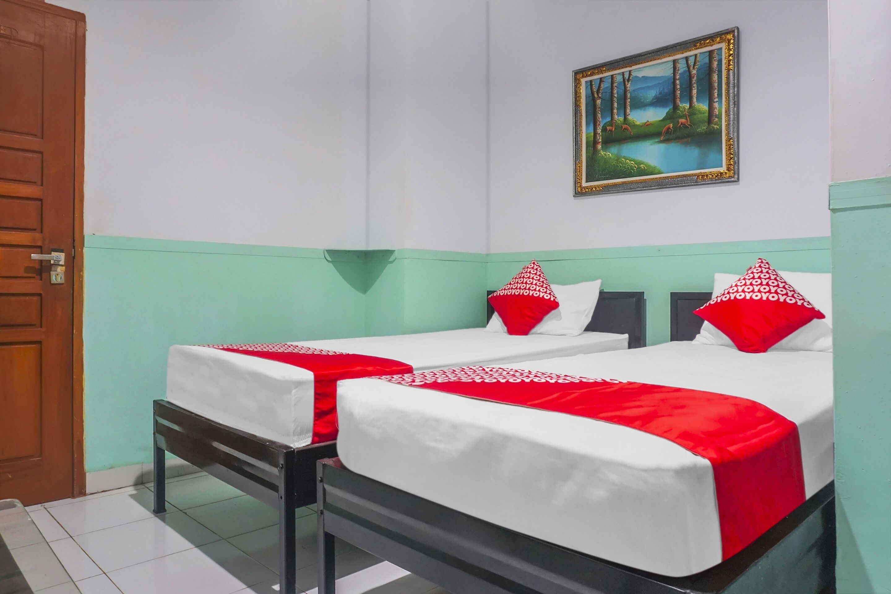 A hotel room with two beds, each with white bedding and red pillows, a wooden door, and a painting of a forest scene on the wall.