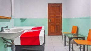 A bathroom with a sink, two beds with red and white bedding, a wooden door, and a table with orange chairs.