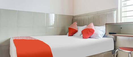 Una cama con una colcha blanca y roja, dos almohadas rojas y un cabecero gris en una habitación con piso de baldosas y una ventana.