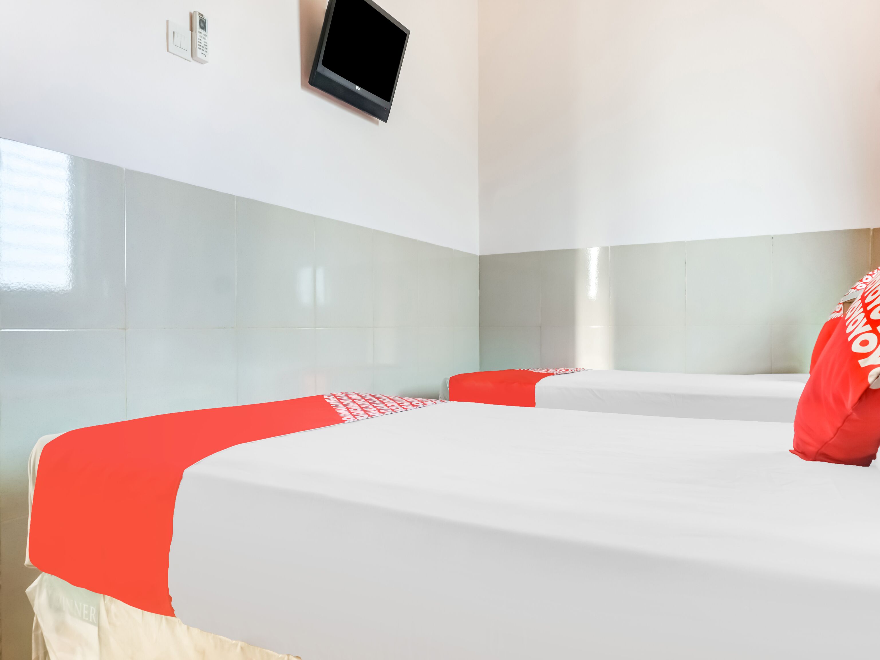A hotel room with two beds, each with a red and white bedspread, and a flat-screen TV mounted on the wall.