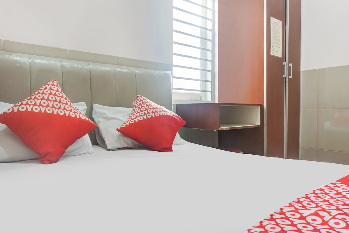 A hotel room with a bed, two red and white patterned pillows, a wooden wardrobe, and a window with blinds.