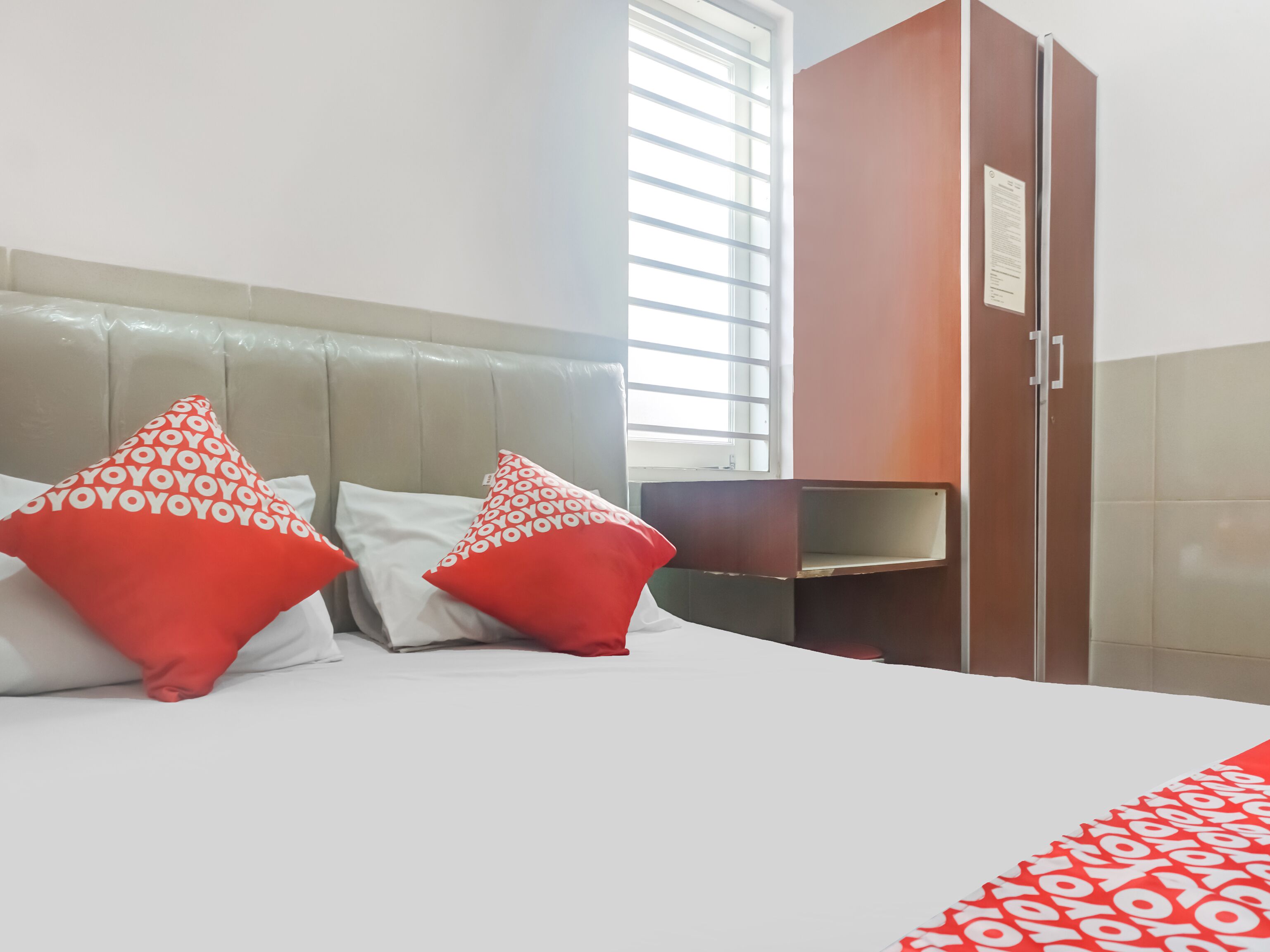 A hotel room with a bed, two red and white patterned pillows, a wooden wardrobe, and a window with blinds.
