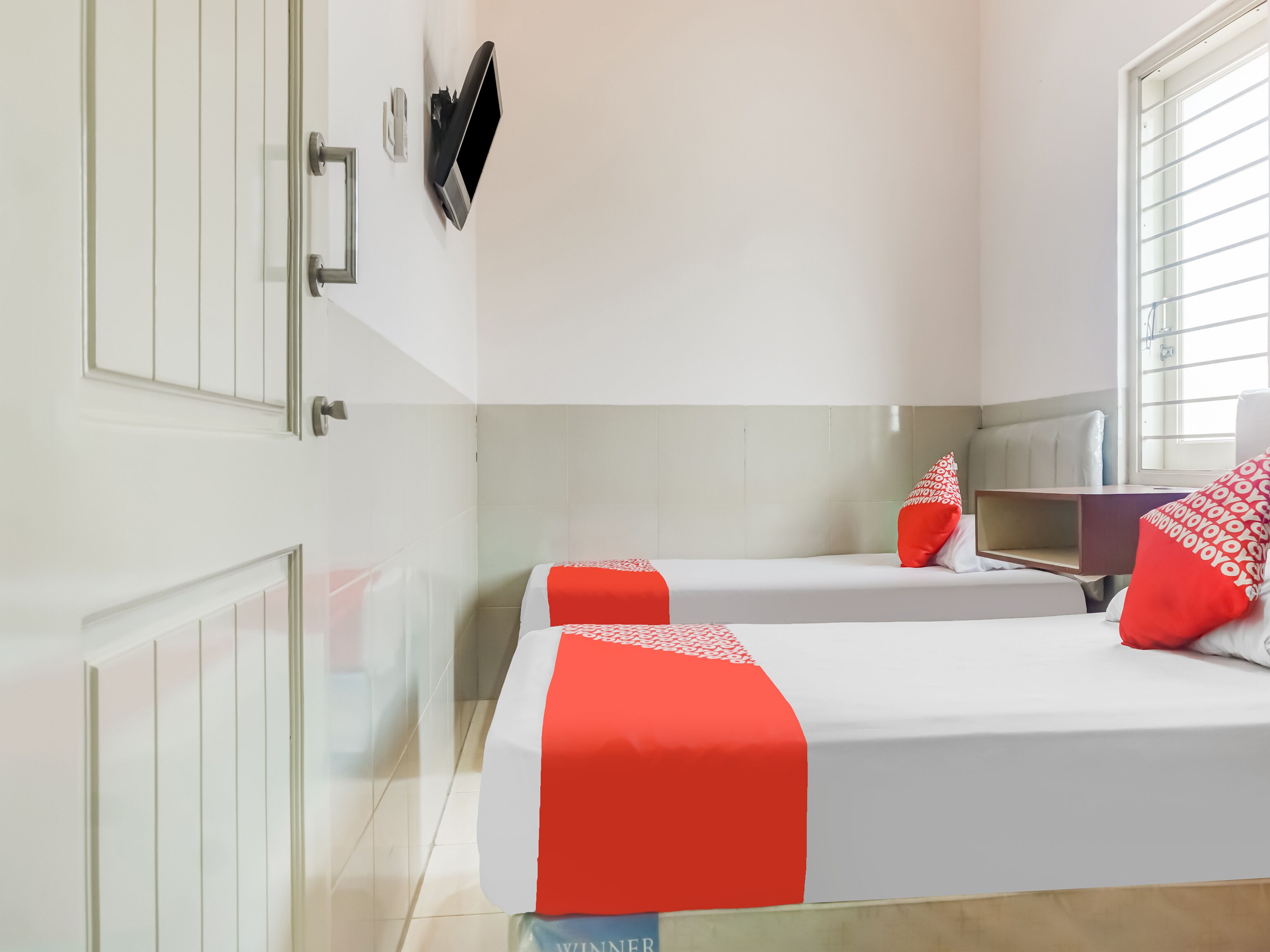 Une chambre d’hôtel avec deux lits, un téléviseur fixé au mur, une fenêtre avec des stores et une table de chevet avec une lampe.