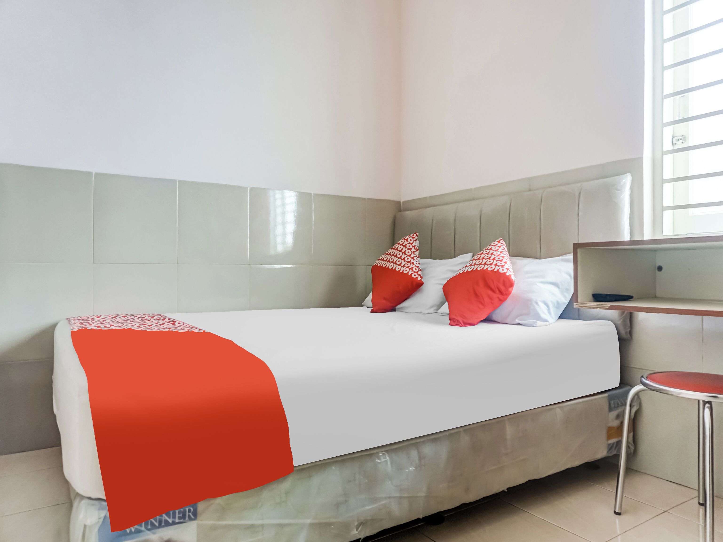 A bed with a white and red bedspread, two red pillows, and a grey headboard in a room with a tiled floor and a window.