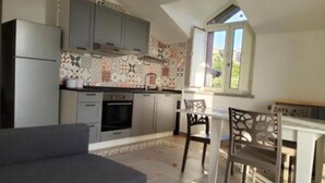 Uma cozinha compacta com armários cinzentos, forno embutido e painel de fundo padronizado. Há uma área de refeições com mesa e cadeiras, e uma janela que oferece luz natural.