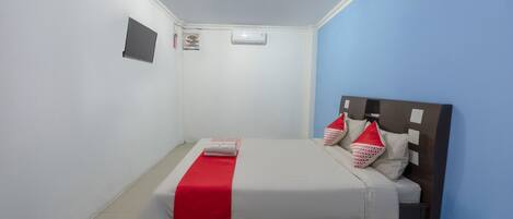 A single bed with a red and white bedspread, a headboard with multiple pillows, and a small air conditioning unit on the wall.