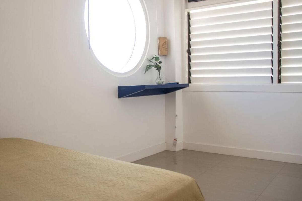 Une chambre avec un lit, une fenêtre avec des stores, une petite étagère avec une plante et un miroir rond.
