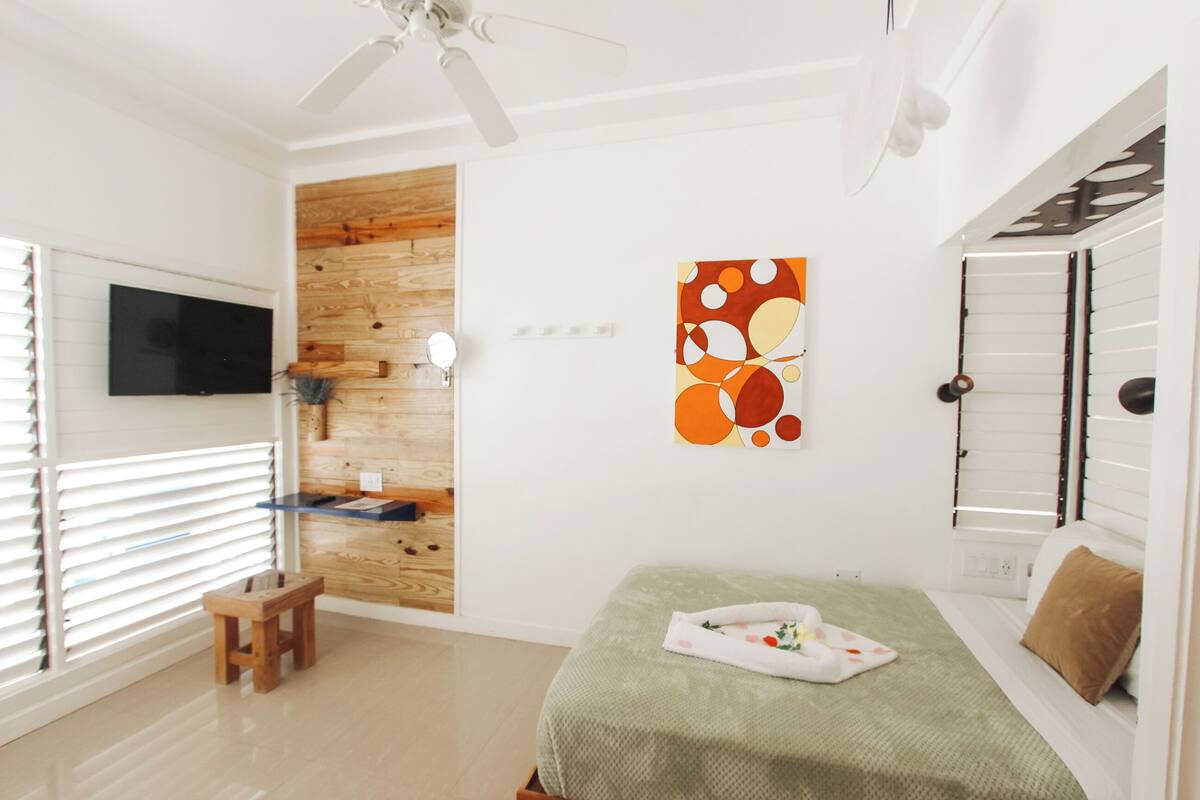 Une chambre à coucher avec un lit, un mur décoratif en bois, un ventilateur de plafond, une télévision et une fenêtre avec des stores.
