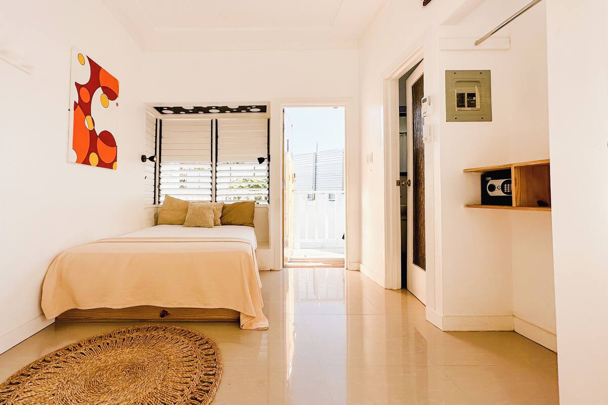 Une chambre à coucher avec un lit, un tapis tissé, une porte avec un trou de serrure et un mur orné d’un tableau aux motifs géométriques.