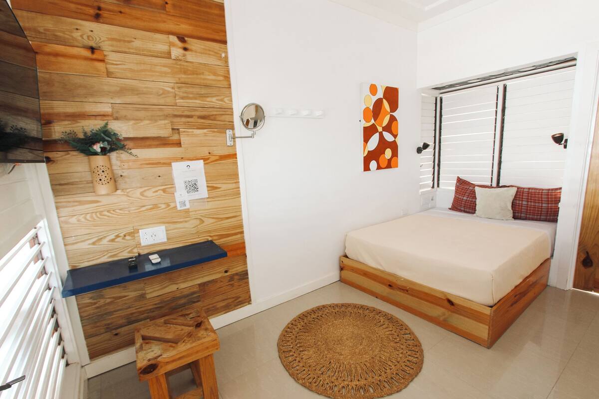 Une chambre avec un mur en bois, un lit avec un matelas blanc, une petite table en bois, un tapis rond tissé et un luminaire fixé au mur.