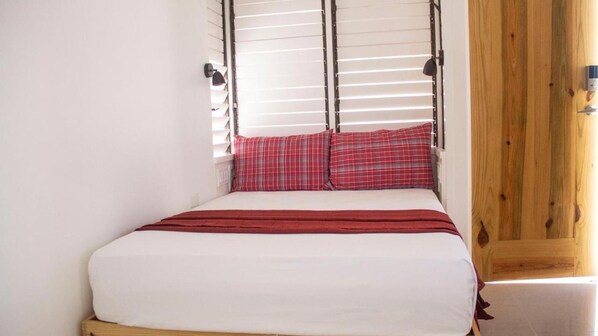 A bed with white bedding and red plaid pillows, a wooden headboard, and a window with blinds.