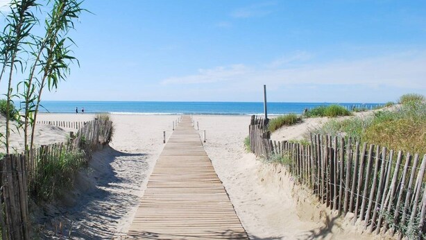 Ein hölzerner Steg führt zu einem Sandstrand bei blauem Himmel.