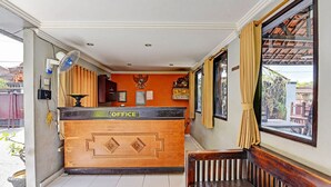 Une salle d’attente avec un comptoir en bois portant l’inscription « BUREAU », un ventilateur et une fenêtre avec des rideaux.