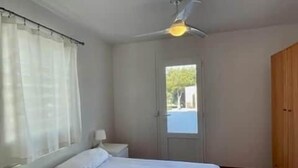 Une chambre à coucher avec un lit, un ventilateur de plafond, une fenêtre avec des rideaux, une porte et une armoire en bois.