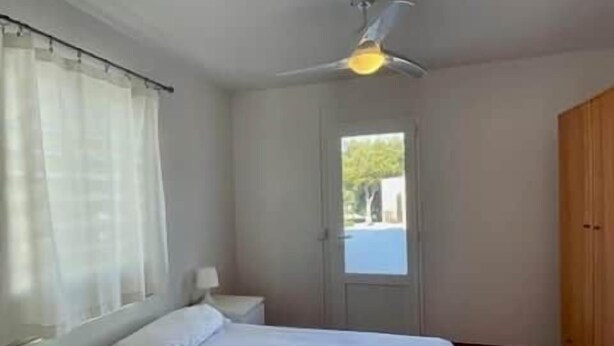 Un dormitorio con cama, ventilador de techo, ventana con cortinas, puerta y armario de madera.