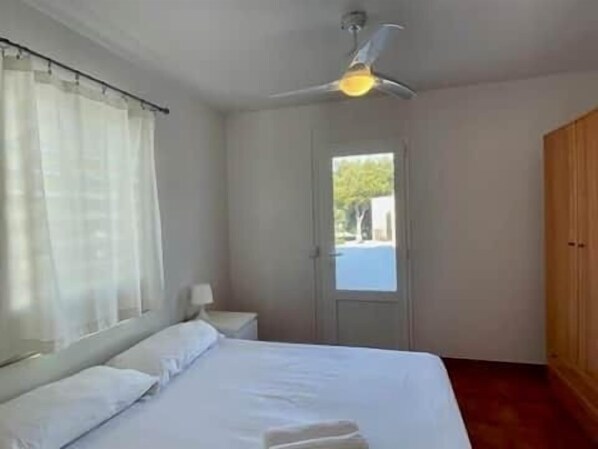 A bedroom with a bed, a ceiling fan, a window with curtains, a door, and a wooden wardrobe. - Apto. S'aigua dolça 3 (Son Carrió)