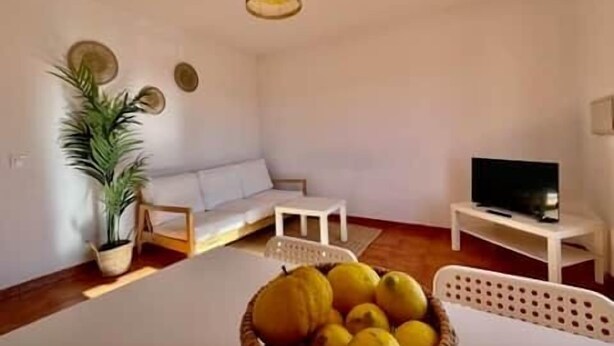 Un salon avec un canapé blanc, un téléviseur posé sur un meuble, une table à manger agrémentée d’un panier de citrons et un luminaire suspendu.