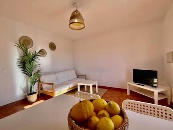 Un salon avec un canapé blanc, un téléviseur posé sur un meuble, une table à manger agrémentée d’un panier de citrons et un luminaire suspendu.