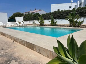 Une piscine rectangulaire bordée de béton, entourée de murs blancs et d’une haie.
