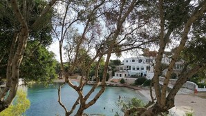 Vista costera con un mar azul cristalino, un edificio blanco con varios balcones y un árbol en primer plano.