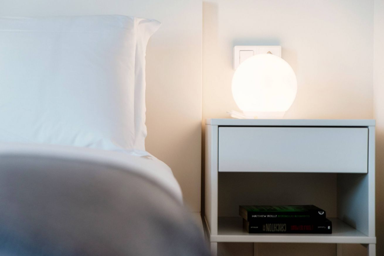 A neatly made bed with white linens, a white nightstand with a lamp, and a few books on it.