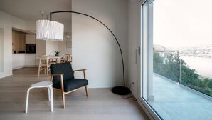 A modern living room with a curved black floor lamp, a dark grey armchair, a white side table, and a view of a cityscape through large windows.