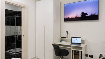 Un dormitorio moderno con ducha envuelta en vidrio, cama, escritorio con una computadora portátil y televisor mostrando una escena de atardecer.