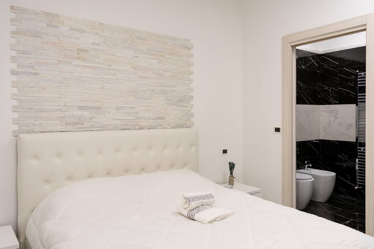 A bedroom with a white bed, a tufted headboard, a stone wall, and a bathroom with black marble tiles.