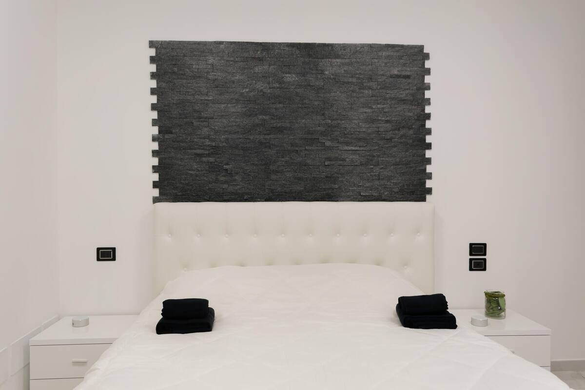 A bedroom with a white bed, a grey textured headboard, a white nightstand, and a small green vase on the nightstand.