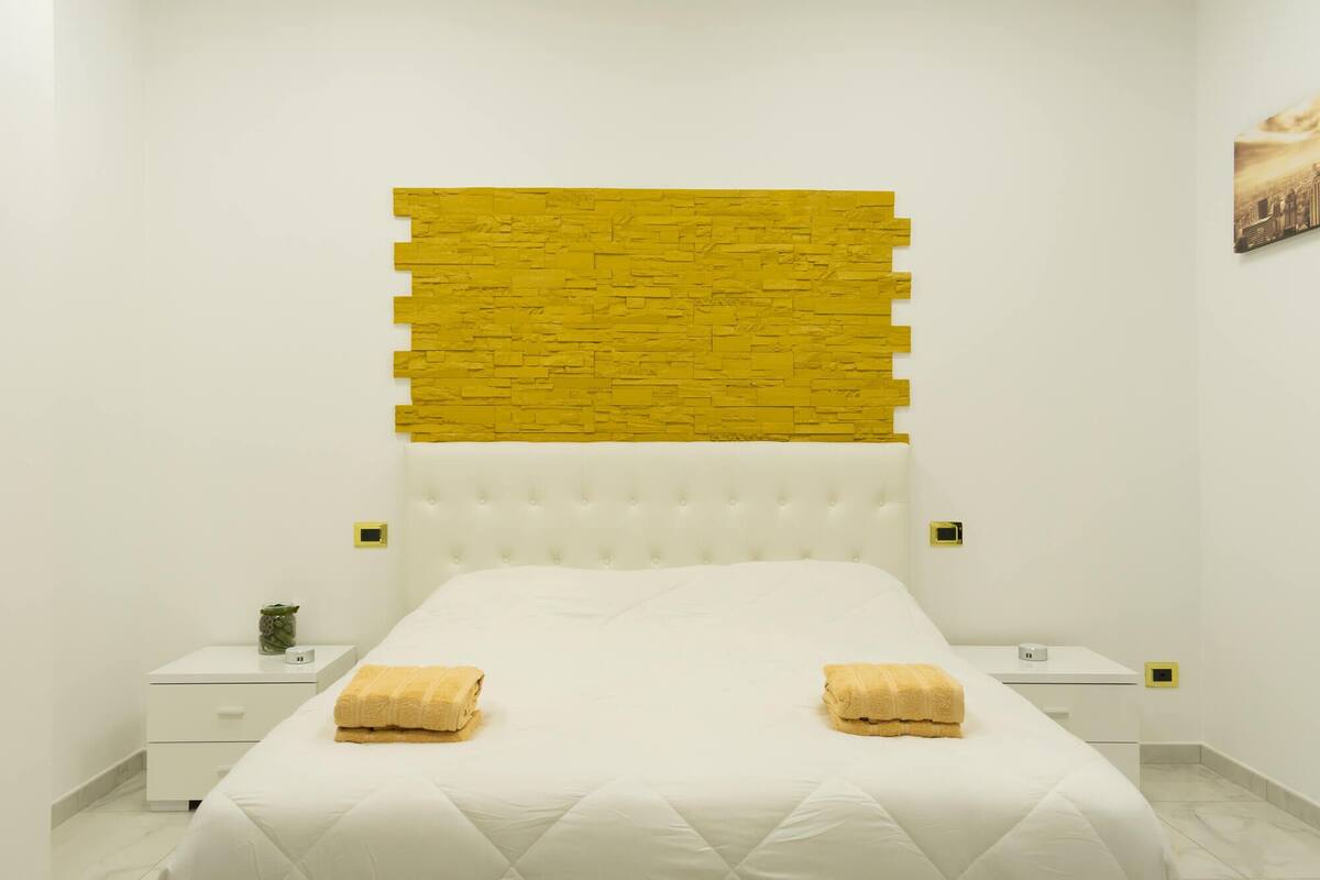 A hotel room with a white bed, a yellow textured wall, and a white nightstand with a small plant.