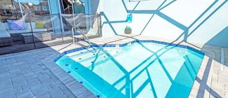 Una piscina con cerramiento de vidrio, un área de descanso con sofá y sillones, y vista a un edificio con grandes ventanales.