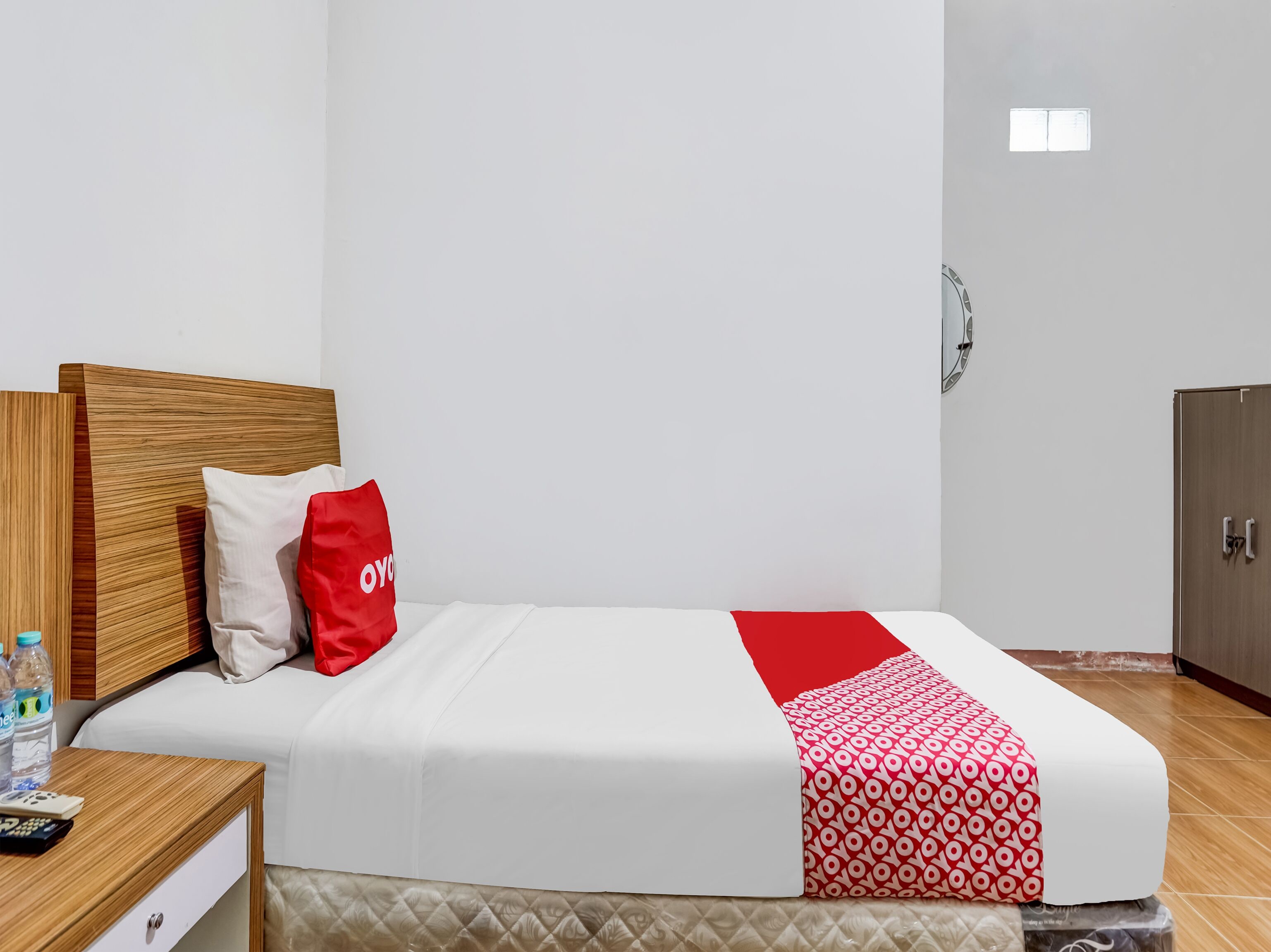 A hotel room with a bed, a wooden headboard, a red and white patterned blanket, a white pillow, and a wooden nightstand with a remote control and water bottles.