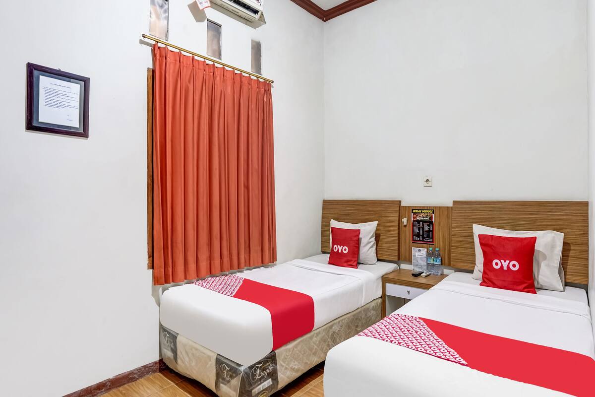 Une chambre d’hôtel avec deux lits, chacun recouvert d’une courtepointe rouge et blanche, et agrémentée d’oreillers assortis. On y trouve une tête de lit en bois, une table de chevet avec un téléphone et des bouteilles d’eau, et un certificat encadré accroché au mur.