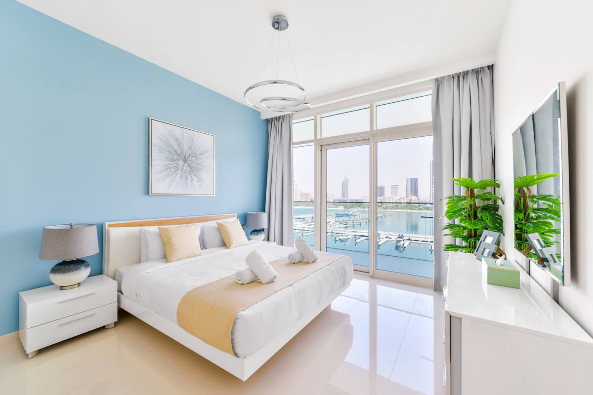 A modern bedroom with a large bed, a nightstand, a lamp, a view of the city through large windows, and a potted plant.