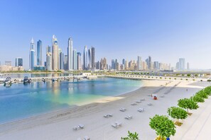 Un paysage urbain côtier avec des gratte-ciel modernes, une marina remplie de bateaux et une plage avec des transats.