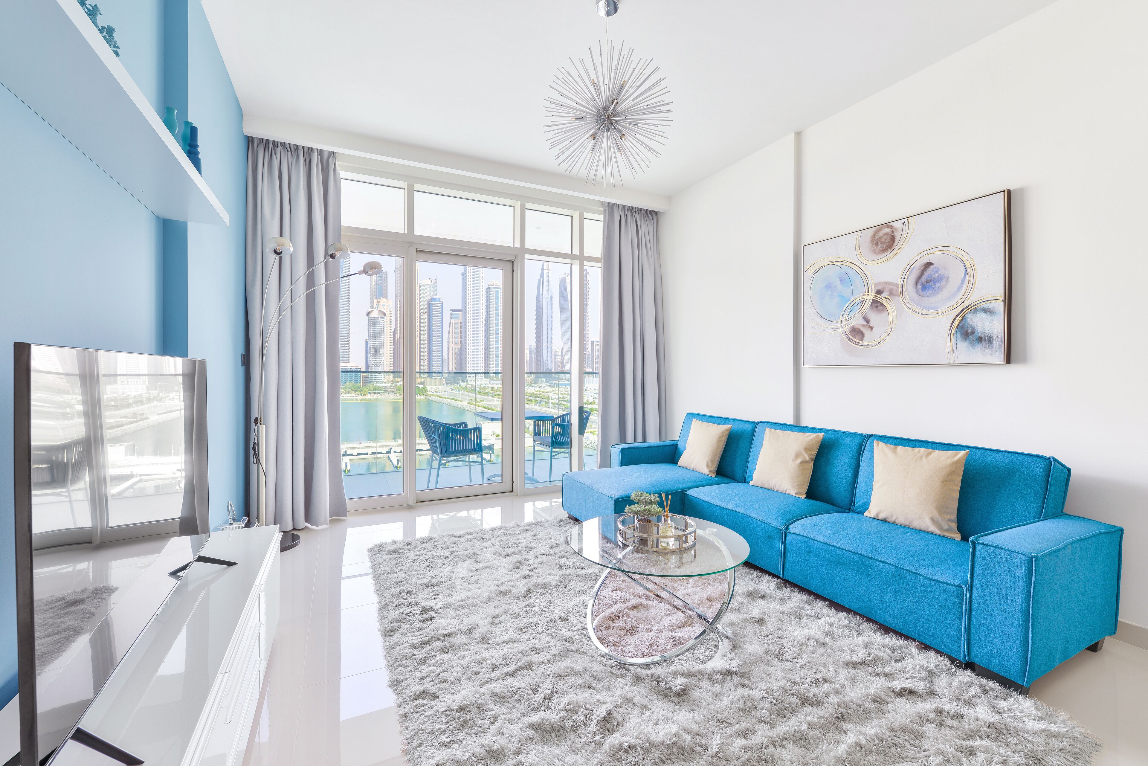 Uma sala de estar moderna com um sofá azul, uma mesa de centro de vidro e vista para a cidade através de grandes janelas.