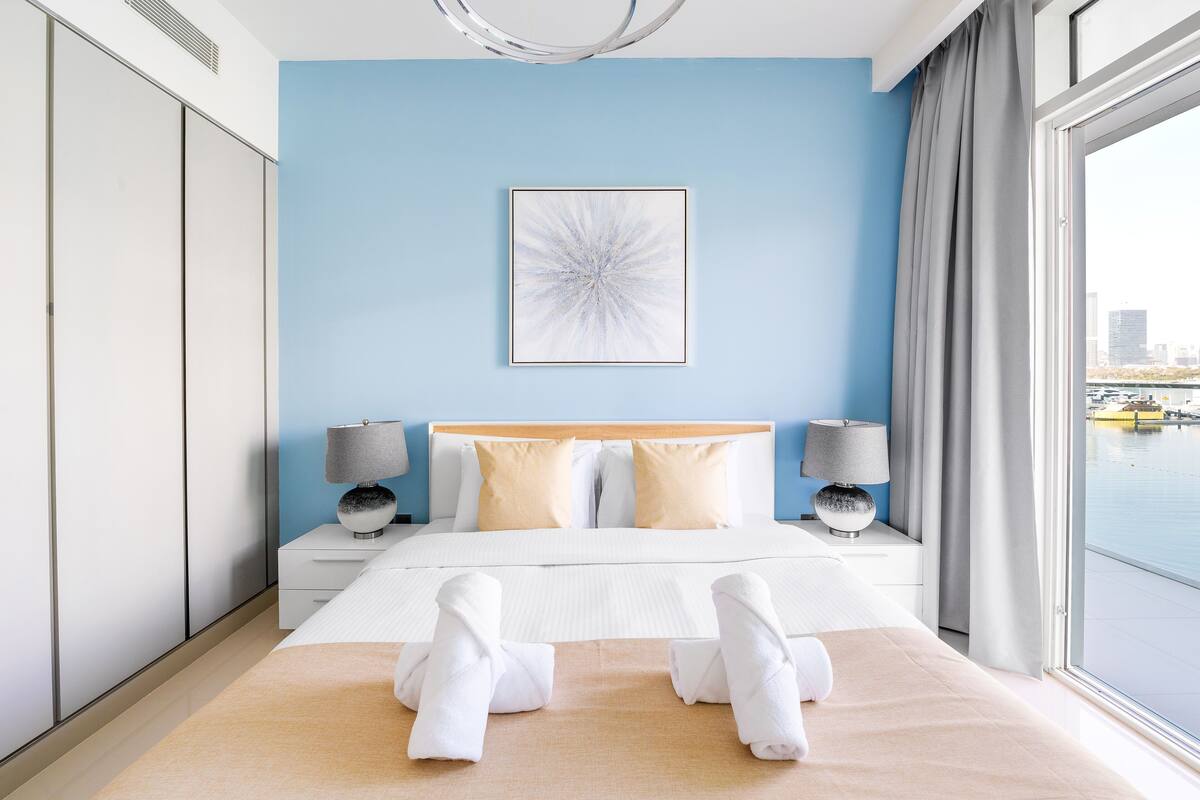A modern hotel room with a large bed, two bedside tables with lamps, a city view, and a blue accent wall.