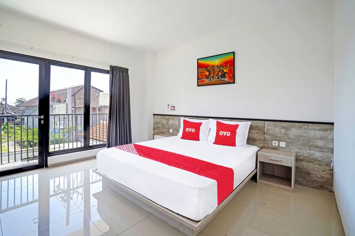 A hotel room with a large bed, a nightstand, a painting on the wall, and a view through a sliding door.