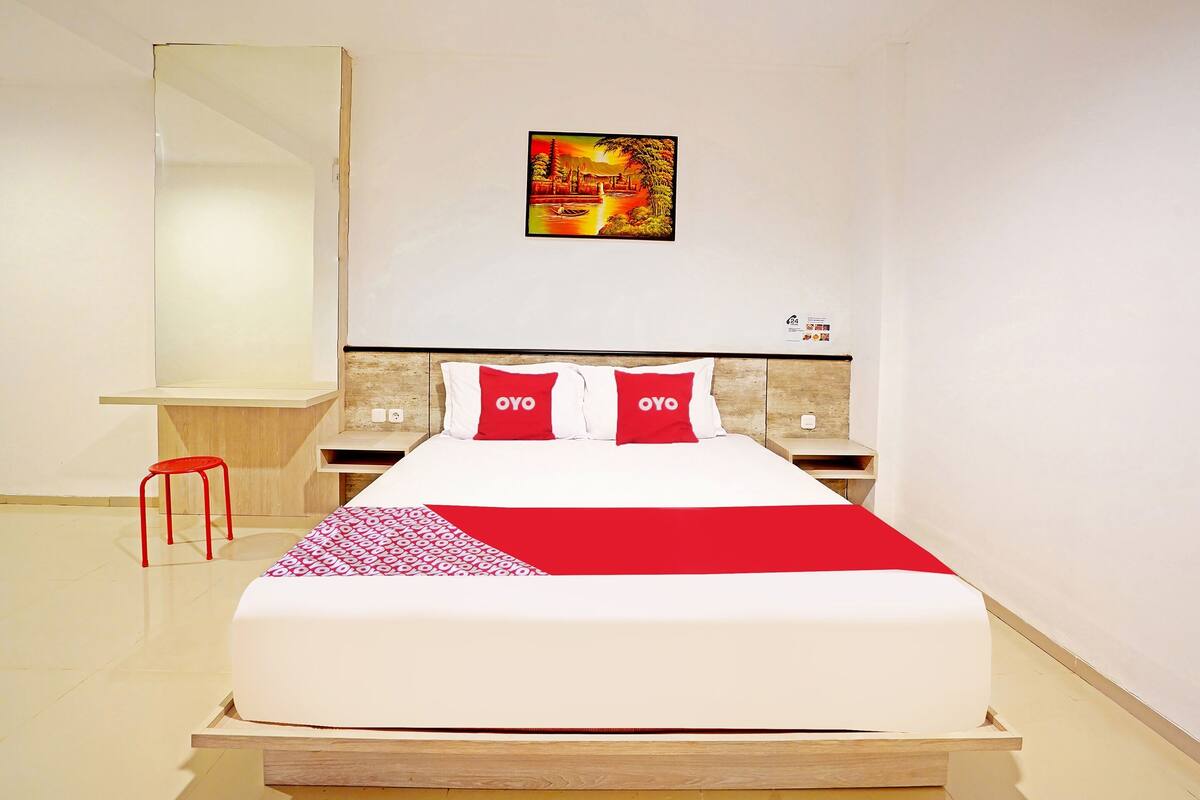 A modern hotel room with a large bed, a red and white bedspread, two red pillows, a nightstand with a lamp, and a wall-mounted painting.