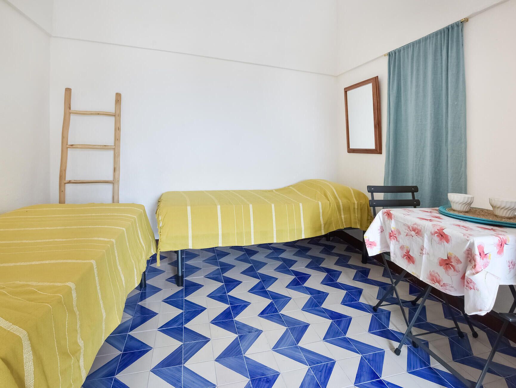 Una habitación con dos camas, una mesa con mantel floral, una silla, una escalera y un espejo en la pared.