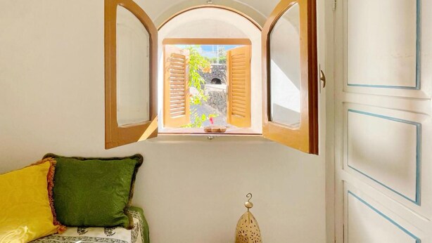 Ein Zimmer mit Fenster, Holzläden, einem grünen Kissen und einem dekorativen Gegenstand auf dem Boden.