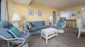 A living room with a blue sofa, a wicker coffee table, and a chair with a striped cushion. There is a kitchen visible in the background.