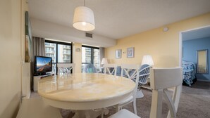 A hotel room with a round dining table, white chairs, a television, and a large window with a view of buildings.