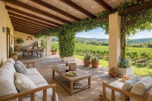An outdoor patio with a wooden ceiling, cushioned seating, a table with wine glasses, and potted plants.