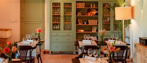 Un coin repas avec des tables préparées pour deux personnes, chacune agrémentée d’un vase de fleurs, de verres et de couverts. Au fond, on aperçoit des meubles verts avec des portes en verre.