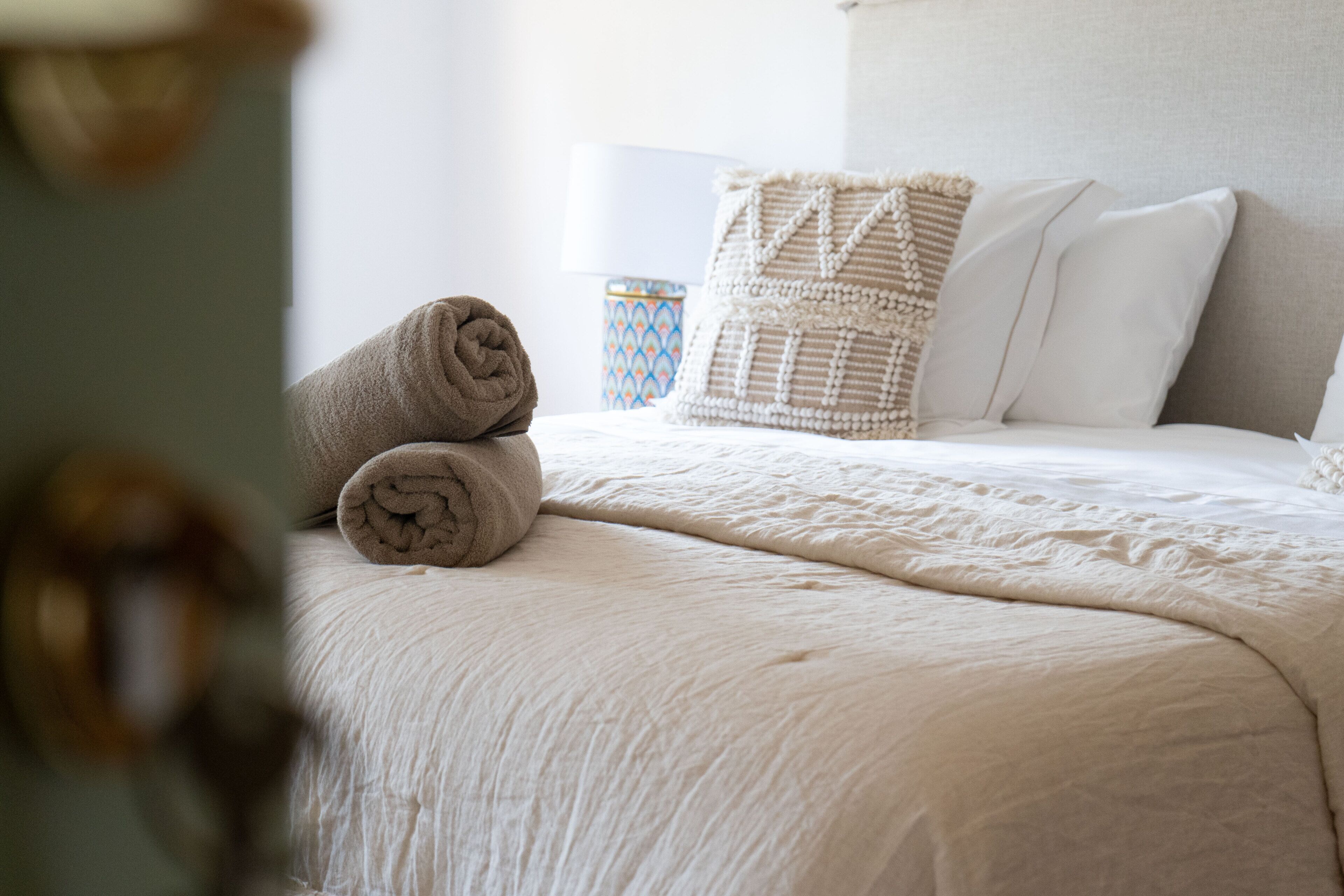 Una cama bien hecha con un edredón blanco, almohadas, una toalla enrollada y una almohada decorativa estampada.