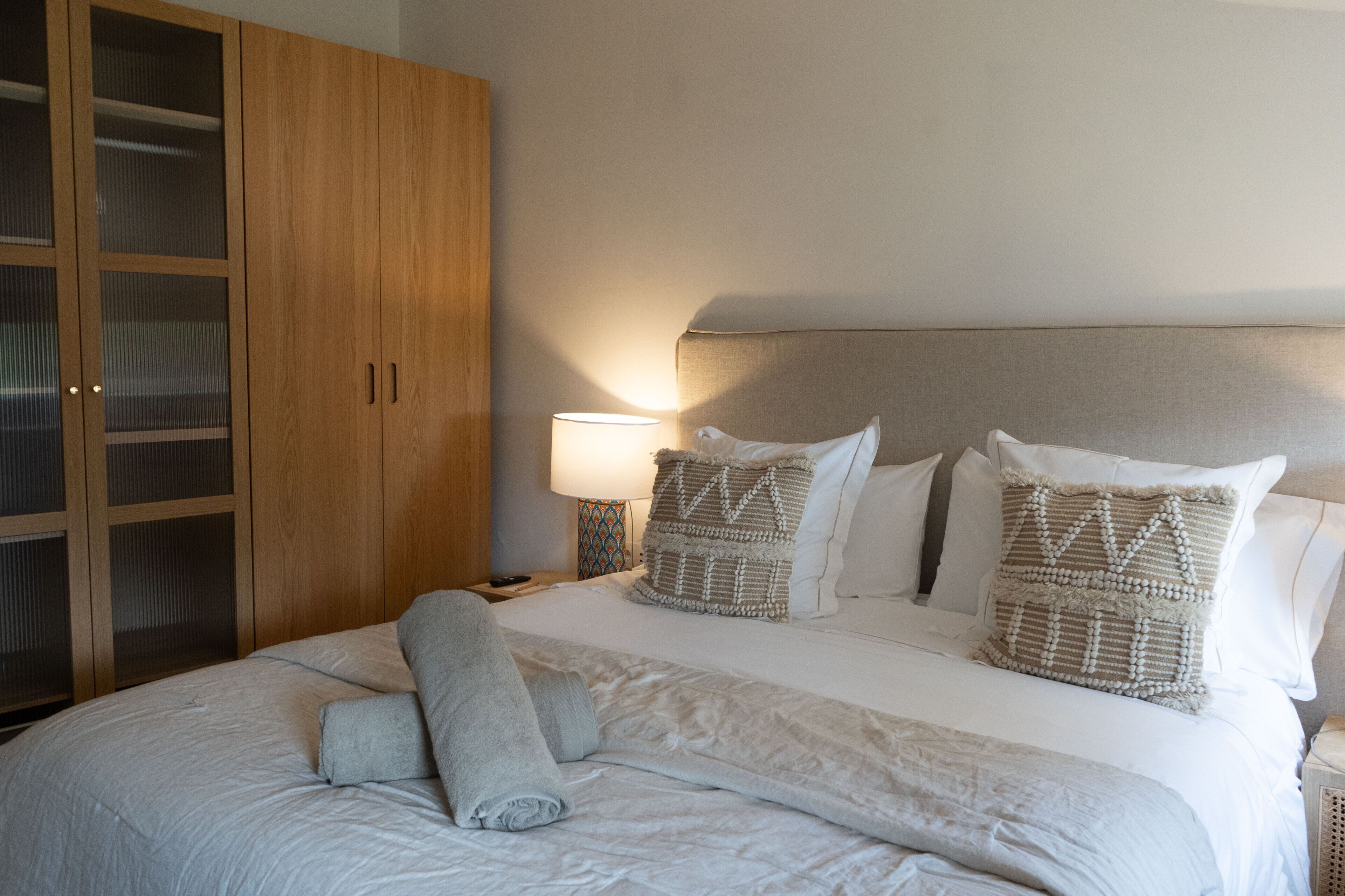A neatly made bed with a rolled towel, a wooden wardrobe, and a bedside lamp.