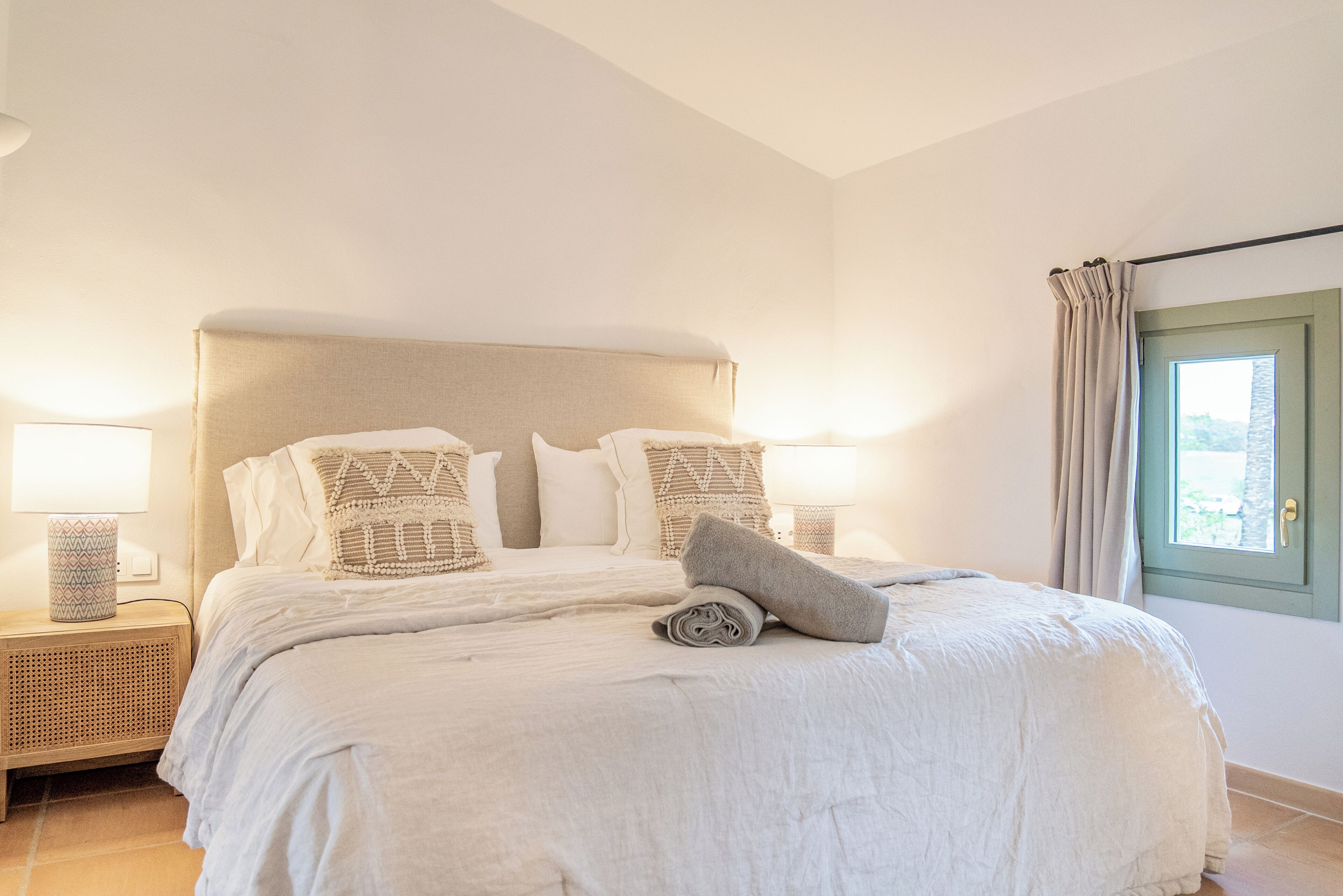 Una cama bien hecha con cabecera, dos lámparas de noche y una ventana con cortinas.